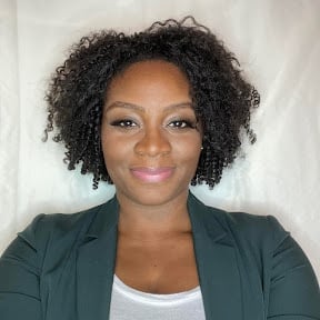 Professional headshot of a woman with dark curly hair wearing a dark green blazer and white top against a light background