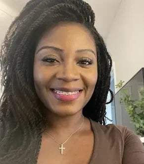 Woman with long dark braided hair wearing a brown top and silver cross necklace, smiling at the camera indoors
